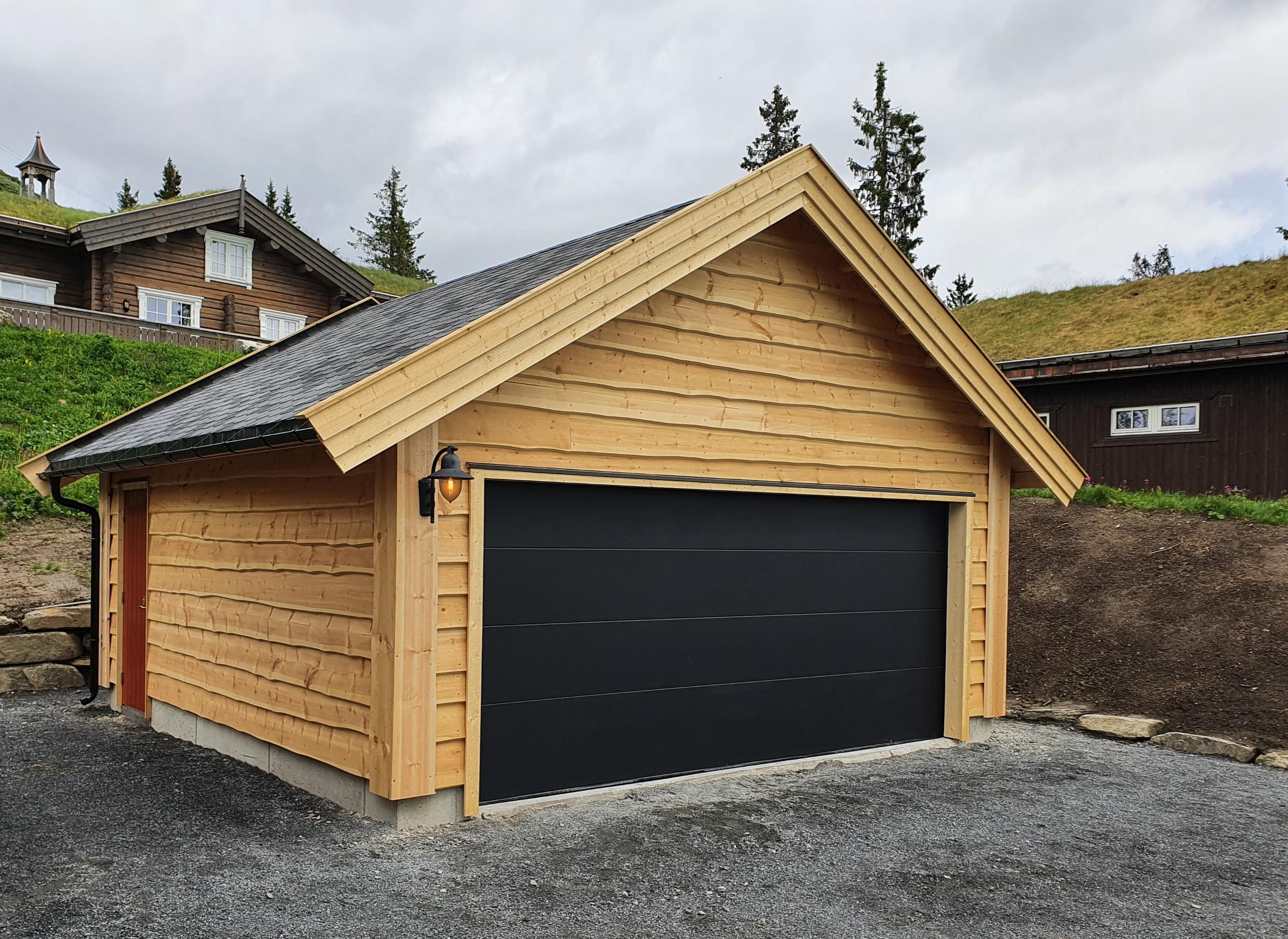 Ny garasje i lys trepanel med svart port, torvtak på hyttene bak