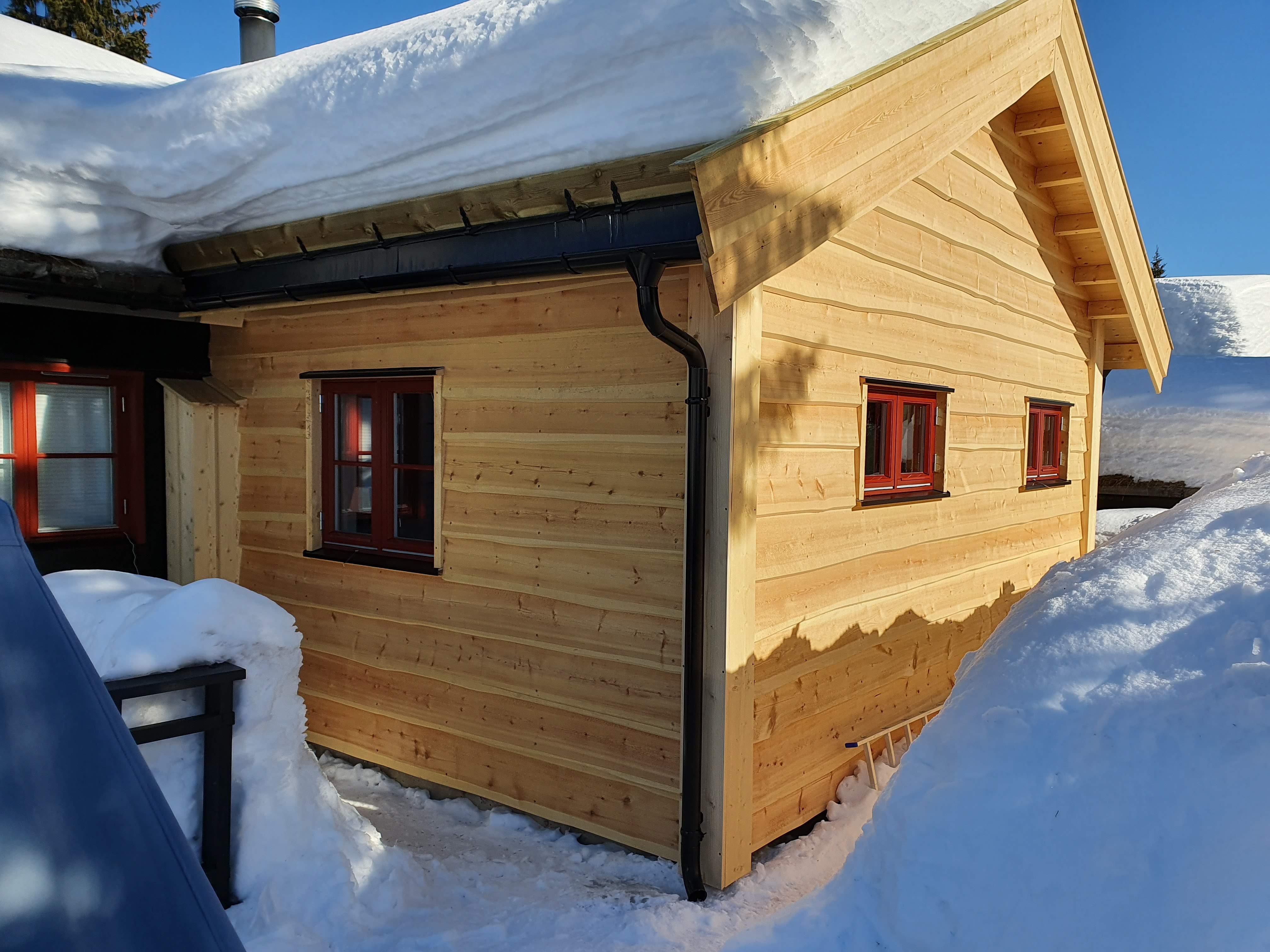 Hyttevegg i tre med røde vindusrammer, snø til under vinduene